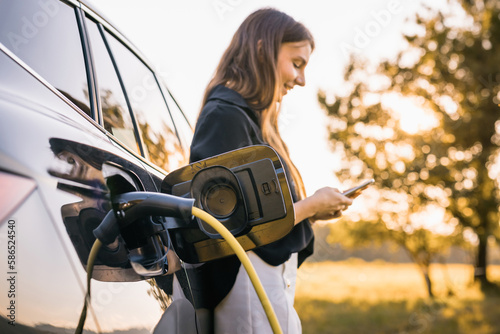 Girl charging electric car parked in the nature area and adjusting an EV charging app on smartphone