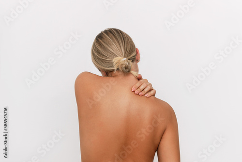 Young tanned blonde woman standing with her back holds her neck massaging a trapezius muscle with her hand isolated on a white background. The girl suffers from an osteochondrosis