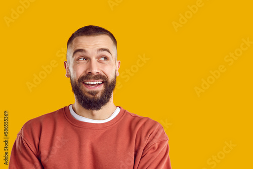 Portrait of happy tricky smiling bearded brunet man in red sweatshirt looking aside on copy space on yellow background. Banner for advertisement, marketing with good offer. Sincere emotions concept.