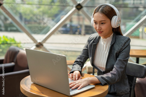 Young asian businesswoman working on her computer laptop and wearing a white color headphone in a cafe. Beautiful lady listen to music while working. Hybrid workplace.