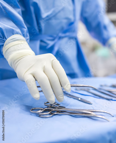 Hand of doctor or surgeon in blue gown pick up surgical clamp instrument or tools inside operating theatre with blur background.Medical equipment for surgery inside room in hospital.Modern technology.