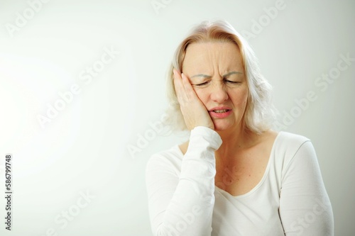 An adult woman has a toothache, she presses her hand to her cheek Toothache blonde woman in all white stands against the background in clinic dentist Health disorders waiting for the doctor