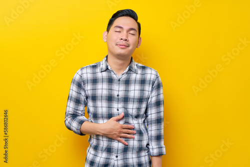 Young Asian man wearing plaid shirt holding hands on his belly suffering from stomach pain or hunger isolated on yellow background. people lifestyle concept