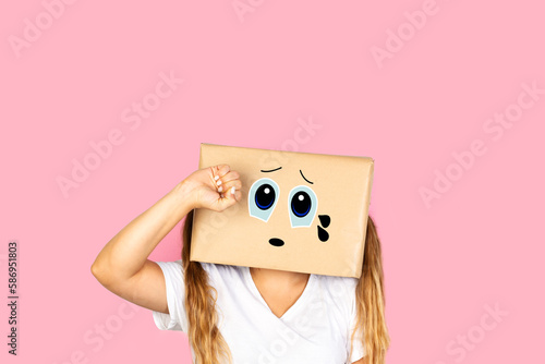 Mujer con caja de cartón en la cabeza con gesto de tristeza y con lágrimas en los ojos sobre fondo rosa liso y aislado. Vista de frente. Copy space 