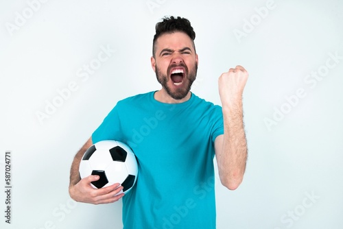 Young man holding a ball over white background angry and mad raising fist frustrated and furious while shouting with anger. Rage and aggressive concept.