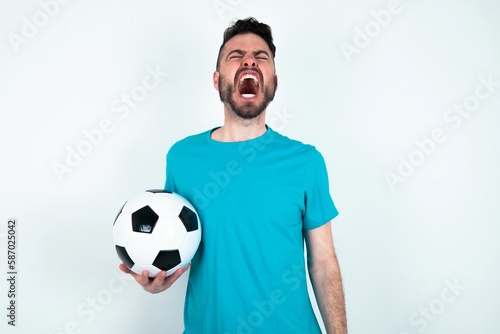 Young man holding a ball over white background angry and mad screaming frustrated and furious, shouting with anger. Rage and aggressive concept.