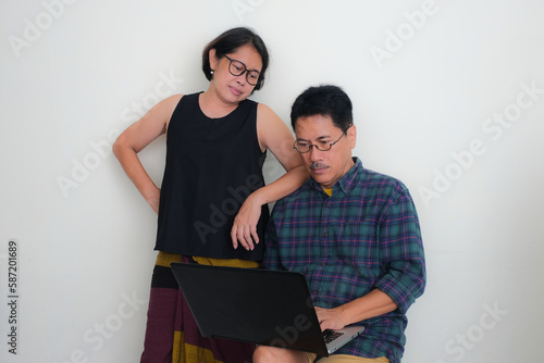 A man working on his laptop, his wife standing nearby, her elbow on her husband's shoulder, both staring at the laptop intensely.