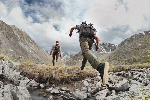 Two young hikers walks with light backpacks in mountains. Tourist jumps across the obstacle