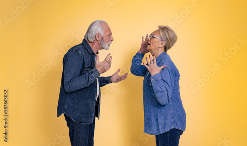 Side view of furious senior couple dressed in blue jeans arguing and screaming at each other while standing face to face isolated over yellow background