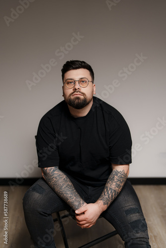 Content young plus size bearded male model in black outfit and eyeglasses with tattooed arms sitting on chair against gray background in studio