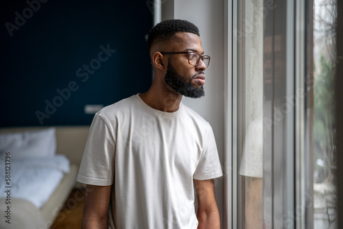 Sad Black man looking out of window thinking about life problems. Lonely African American guy at home dealing with breakup depression. Worried male pondering before making decision. Loneliness concept