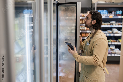 Side view portrait of male worker in supermarket inspecting freezer inventory and holding digital tablet