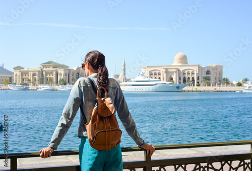 A young woman tourist with a backpack looks at the administrative region of the emirate of Sharjah with the port and ships.
