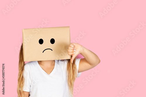 Mujer con caja de cartón en la cabeza enojada y haciendo con el dedo pulgar de la mano un gesto hacia abajo sobre un fondo rosa liso y aislado. Vista de frente y de cerca. Copy space