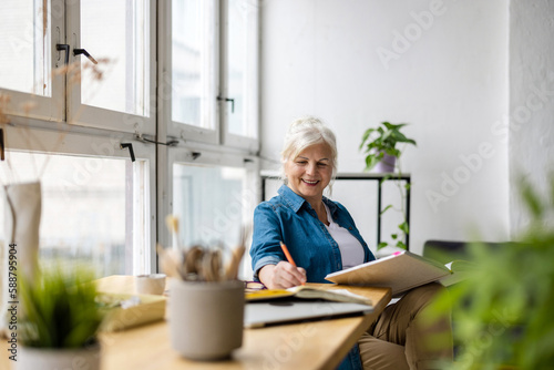 Smiling mature businesswoman writing in notebook while sitting at table in office