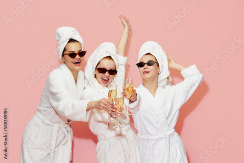 Three beautiful, happy, cheerful girls in bathrobes and towels, drinking champagne, celebrating against pink studio background. Concept of youth, face care, beauty, friendship, hen party, relaxation