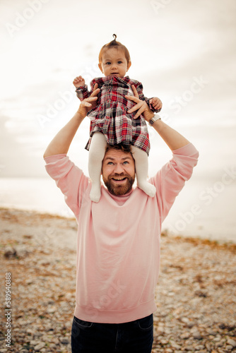 Cute bearded curly daddy smiling playing keeping high at the shoulders his small toddler girl in checkered red dress at Italian Garda lake coast 