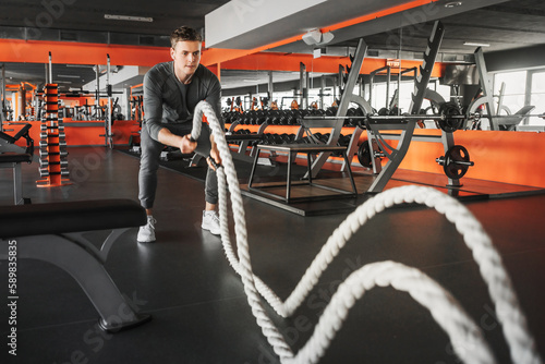 Handsome muscular man is doing battle rope exercise while working out in gym. The guy is dressed in a gray tracksuit and white sneakers. Wide angle shooting.