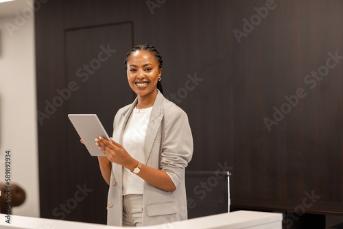 The young African-American female receptionist stands behind the reception desk with a digital tablet in her hands and smiles at the camera.