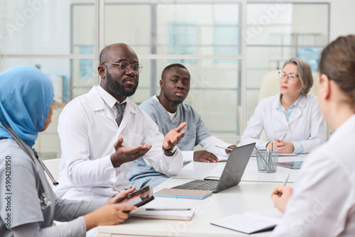 African American doctor in white coat sitting at table with laptop and discussing the way of treatment with other colleagues at meeting