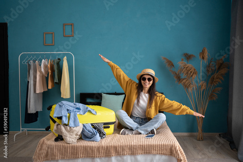 Laughing young woman sitting on bed with suitcase at home, holding hands apart in flight ready for summer vacation. Traveling overseas, plane flight