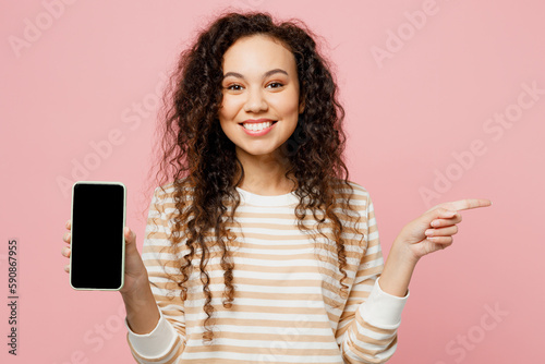 Young woman of African American ethnicity she wear light casual clothes hold in hand use mobile cell phone with blank screen workspace area point finger aside isolated on plain pastel pink background.