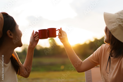 Happy cheerful Asian young women - friends camping in the lake forest and enjoy drinking a cup of tea or coffee. 