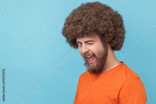 Portrait of flirting man with Afro hairstyle wearing orange T-shirt in good mood, smiling broadly and winking at camera with toothy smile. Indoor studio shot isolated on blue background.