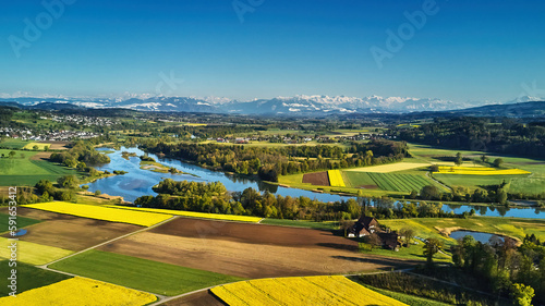 Blick über den Flachsee (Reuss) umgeben von einem bunten Mosaik aus Wald, Wiesen, Rapsfeldern und Ackerflächen, die sich bis zu den Bergen hinziehen. Wolkenloser, stahlblauer Himmel..