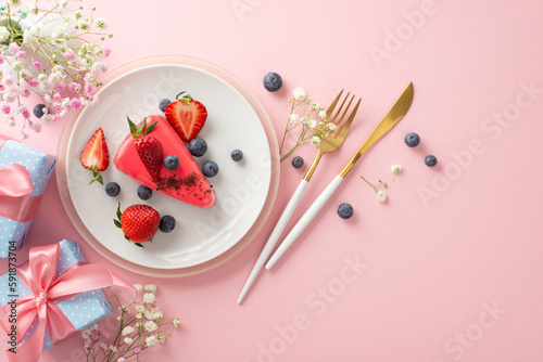 Delicate Mother's day table concept. Top view flat lay of plates, cutlery, tulips, gift box, and piece of tasty cake with berries on pastel pink background
