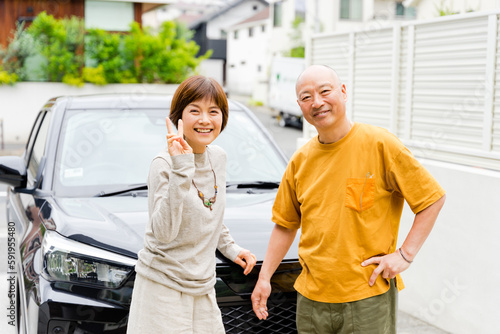 ドライブ前に車の前でピースサインをする50代から60代の仲の良い老夫婦2人