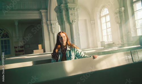 A beautiful young red-haired woman is sitting on old pews inside a Protestant church, illuminated by the radiance of the sun's rays and divine light. Faith and religion.