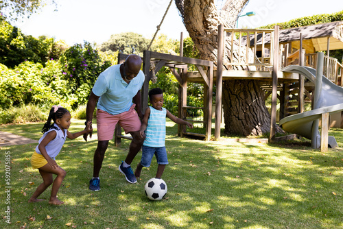 African american grandfather playing soccer with cheerful grandchildren in park