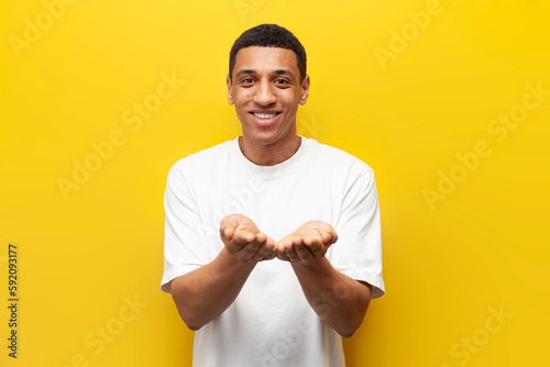 young African American guy holds his hands in front of him on yellow isolated background, man holds nothing