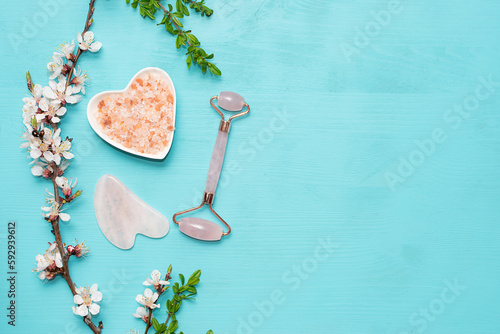 Pink Himalaya salt, Gua Sha massage quartz roller and aromatic blooming apricot branches on bright blue background with copy space for your design. Skin care concept. Spa and wellnes background.