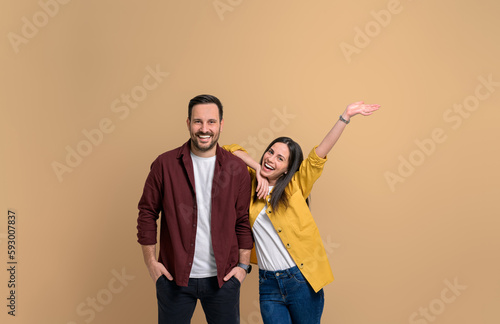 Cheerful young girlfriend with hand raised screaming and leaning on handsome boyfriend. Portrait of attractive couple dressed in shirts posing happily over beige background