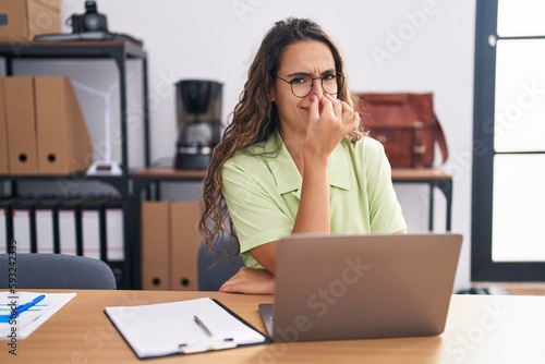 Young hispanic woman working at the office wearing glasses smelling something stinky and disgusting, intolerable smell, holding breath with fingers on nose. bad smell