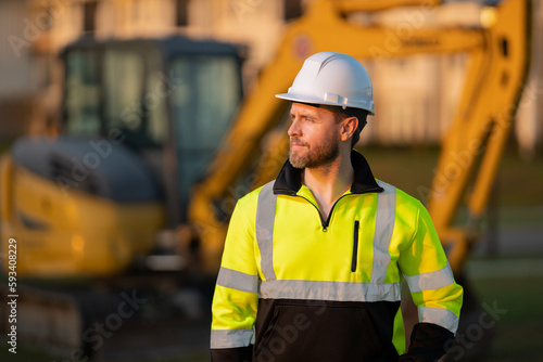 Worker in helmet on site construction. Excavator bulldozer male worker. Construction driver worker with excavator on the background. Construction worker with tractor or construction at building.