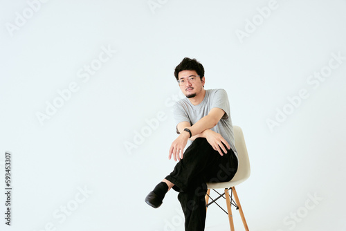 屋内の白バックで1人でおしゃれな椅子に座る30代のカメラ目線の日本人男性