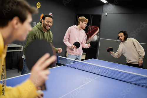 Excited interracial men standing near friends playing table tennis in gaming club.