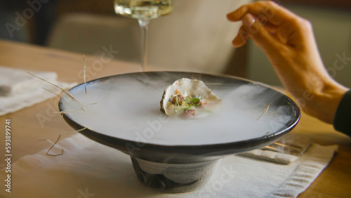 Close up shot of pouring liquid in meal to make dry ice steam for dish presentation in molecular cuisine restaurant. Hand of woman sitting at the table in modern gastro cafe. Concept of public eating.