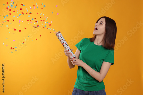 Happy teenage girl blowing up party popper on orange background