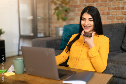 Smiling spanish lady sitting at desk with laptop, talking on speakerphone, dictating voice message, virtual assistant