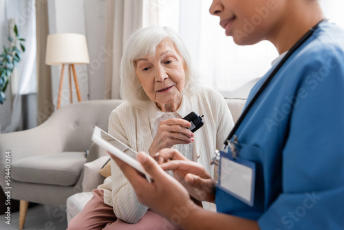 senior woman with grey hair holding glucometer near multiracial nurse with digital tablet.