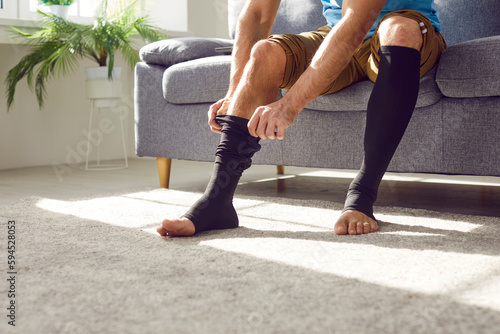 Man wears compression stockings due to varicose or recovery after thrombosis. Unrecognizable man sitting on sofa and covering tired legs with elastic medical anti varicose anti thrombotic sleeve socks