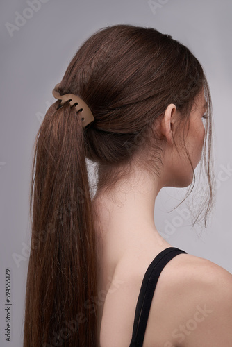 A half-turned brown-haired girl with a ponytail is wearing a black tank top. The lady's hair is fixed with a brown chocolate matte banana hair clip. The photo was taken on a light background.