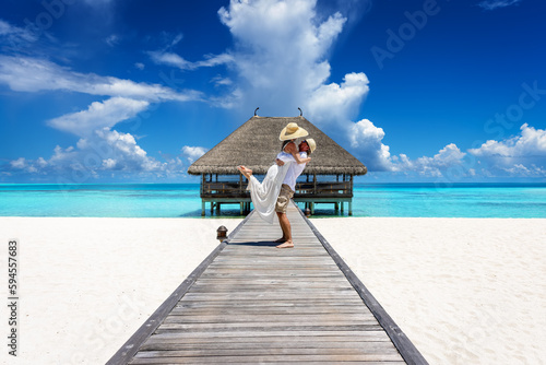 A happy couple in white summer clothing hugging on a wooden pier towards a tropical paradise island in the Maldives, Indian Ocean