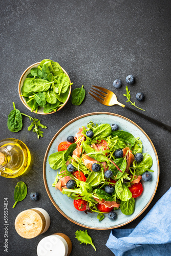 Healthy salad with prosciutto, green leaves mix and tomatoes at dark background. Top view with copy space.