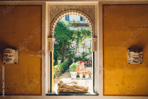 Big window frame with a naked woman statue showing a sunny garden in the Casa de Pilatos palace with masks on the yellow walls.