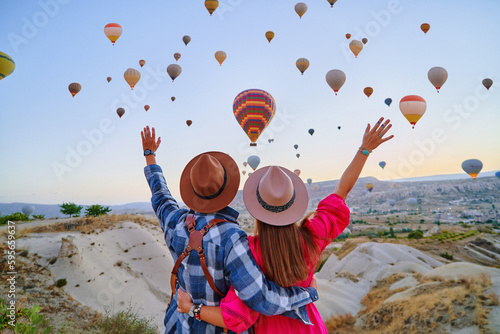 Traveling couple with open arms standing together in scenic valley in Anatolia, Kapadokya. Flying hot air balloons at beautiful destination in Nevsehir, Goreme